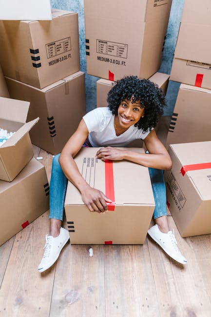 A woman with curly black hair, wearing a white t-shirt and light blue jeans, sitting on a wooden floor surrounded by numerous cardboard boxes of various sizes, many sealed with red packing tape. She is smiling and holding a medium-sized box in her lap, which is also taped securely. The boxes are arranged inside a room with textured, light blue walls, and some are positioned against the wall or on the floor around her. The scene depicts a packing stage in a home relocation or furniture transport process, with visible packing materials and boxes prepared for moving. The image illustrates the packing and loading activities commonly involved in professional removals by companies like Man with Van Maida Vale, supporting efficient and organized house or office moves.