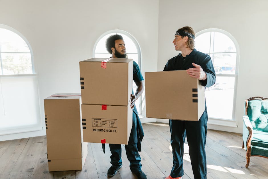 A woman with curly black hair, wearing a white t-shirt and light blue jeans, sitting on a wooden floor surrounded by numerous cardboard boxes of various sizes, many sealed with red packing tape. She is smiling and holding a medium-sized box in her lap, which is also taped securely. The boxes are arranged inside a room with textured, light blue walls, and some are positioned against the wall or on the floor around her. The scene depicts a packing stage in a home relocation or furniture transport process, with visible packing materials and boxes prepared for moving. The image illustrates the packing and loading activities commonly involved in professional removals by companies like Man with Van Maida Vale, supporting efficient and organized house or office moves.