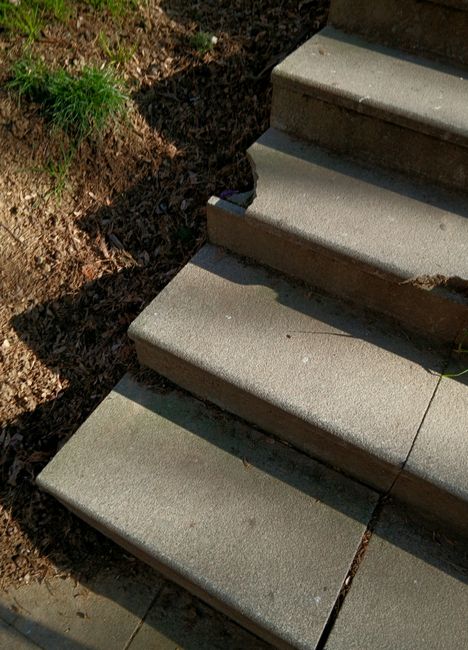 Close-up view of concrete driveway stairs with three steps, situated outdoors in a residential area. The stairs are adjacent to a patch of soil with some grass and small plants on the left side. The steps are light grey with smooth surfaces, slightly rounded edges, and visible lines separating each one, indicating separate slabs. Shadows are cast on the steps, suggesting natural sunlight. The bottom of the stairs is near a paved walkway or driveway area, which is part of the building's entrance. This image illustrates an access point that could present challenges for a home relocation involving furniture transport, requiring careful handling during the loading process. Occasionally, Man with Van Maida Vale has encountered similar outdoor stairways when managing removals in Maida Vale, ensuring safe and efficient movement of household items over such access features.