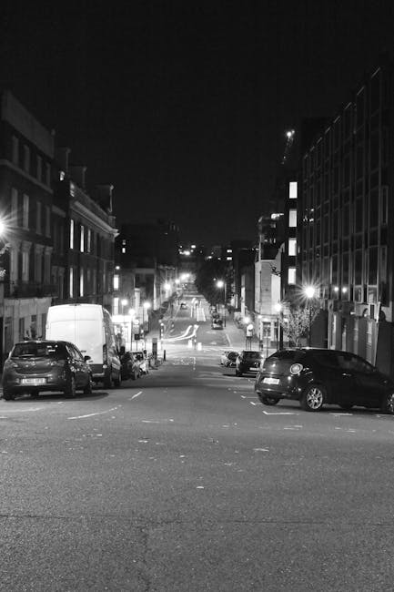 A nighttime street scene in Maida Vale, showing a narrow residential street with parked cars along both sides, including a small hatchback and a larger van. The street is lined with low-rise brick buildings, some with illuminated windows and external lighting. The road surface is dark with visible lane markings, and the area appears quiet with no pedestrians or moving vehicles. In the distance, streetlights and building lights create a subtle glow. This setting illustrates a typical urban environment suitable for house removals or furniture transport, with potential loading or unloading points along the pavement that Man with Van Maida Vale may use for home relocation services.