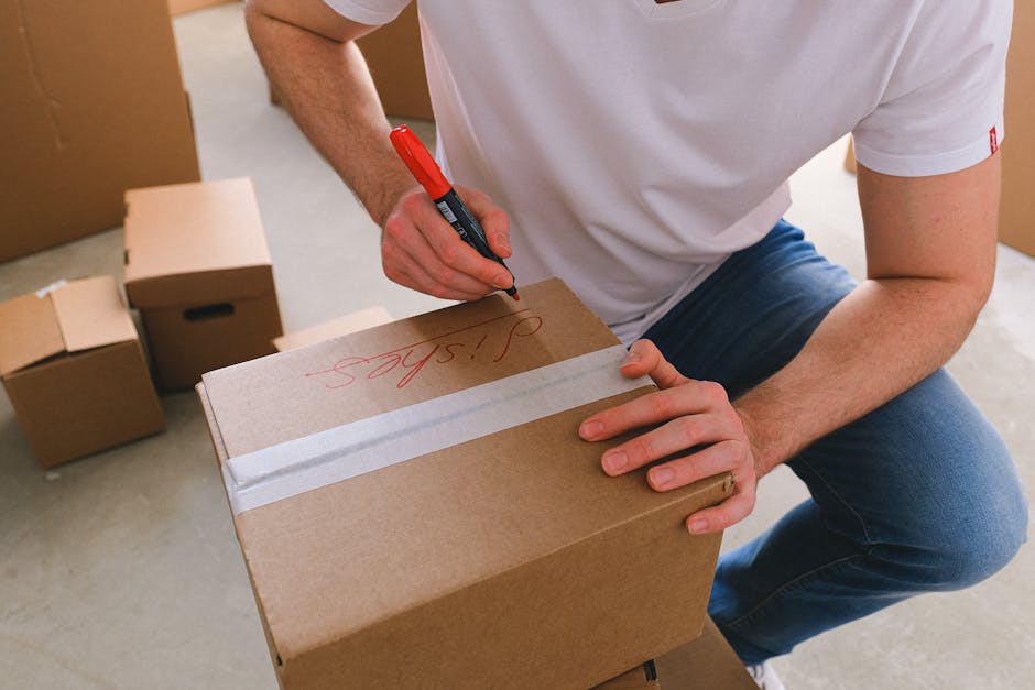 A person wearing a white t-shirt and blue jeans is kneeling indoors, using a red marker to write on the top of a medium-sized cardboard box that is sealed with packing tape. Several other cardboard boxes of various sizes are visible in the background, some of which are open, indicating packing and preparation for removal. The setting appears to be inside a home, possibly a storage or packing area, with neutral flooring and a doorway in the background. This scene illustrates the packing process during house removals, supported by the presence of multiple boxes and packing materials, with natural lighting highlighting the activity. Man with Van Maida Vale specializes in professional removals, including packing, furniture transport, and home relocation services.