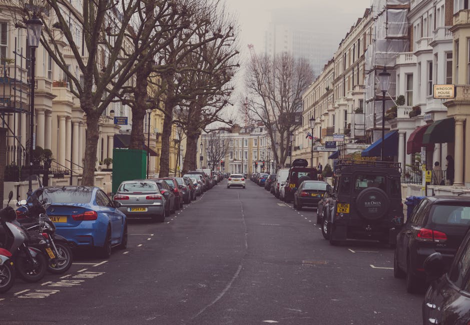 A residential street in Maida Vale lined with parked cars, including sedans, hatchbacks, and SUVs, on both sides of the road. Several motorcycles are parked along the pavement towards the left. Tall, leafless trees with bare branches are situated in front of white Victorian-style terraced houses, which feature bay windows, decorative mouldings, and small front gardens with railings. The sky is overcast, casting diffuse natural light across the scene. In the background, a white van is parked near the curb, while a black off-road vehicle is being loaded with furniture or packed boxes, suggesting a home relocation process. The environment appears to be calm and organized, with no visible pedestrians. The street conveys a typical urban setting suitable for residential removals, with moving equipment such as trolleys or straps possibly used outside the property, supporting the professional activities of Man with Van Maida Vale in their removals service.