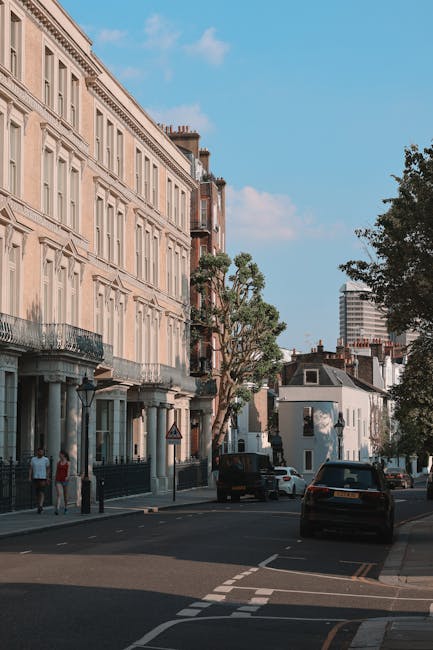 A street scene in Maida Vale showing a row of classic Victorian-style residential buildings with light-colored facades and decorative window frames, situated close to the pavement. Several large cardboard boxes and wrapped furniture items are placed on the sidewalk, suggesting preparations for home relocation. In the background, a black van belonging to Man with Van Maida Vale is parked partially on the road, where other vehicles are also visible, including a dark car parked along the curb. The loading process appears to be underway with individuals possibly engaged in packing or moving activities, although no people are actively seen lifting or carrying objects in this image. The surrounding environment includes a few trees lining the street, a bright blue sky with scattered clouds, and modern high-rise buildings further in the distance, indicating an urban area. The scene captures the typical setting for furniture transport and packing during house removals in Maida Vale, illustrating the logistical aspects of home relocation services.