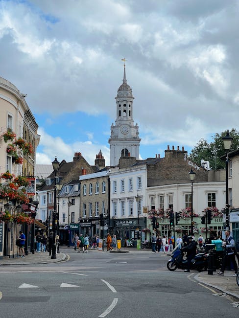 A busy street scene in Maida Vale featuring a historic clock tower with a gold weathervane at the top, situated behind a row of multi-story buildings with a mix of Victorian and Georgian architectural