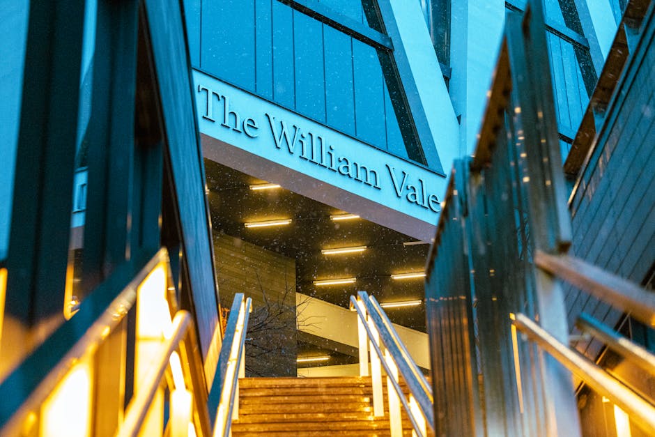 Exterior view of The William Vale building featuring illuminated signage at dusk, with metal and glass building facades visible. The entrance area is approached via a staircase with wooden steps and metal handrails. The surrounding environment includes dark, modern architectural elements, and the lighting creates a warm glow against the darkening sky. This setting relates to home relocation and furniture transport services provided by Man with Van Maida Vale, supporting quick and efficient moving operations such as packing, loading, and transport into or out of this prominent property for house removals.