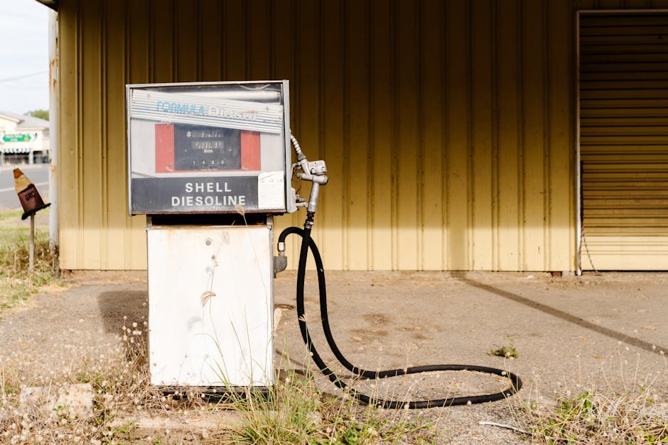A vintage Shell petrol pump, labeled 'Shell Dieseloline,' mounted on a white metal pedestal outside a building with beige vertical siding and a partially open roller shutter door. The pump features a metal hose with a handheld nozzle hanging from its side and is situated on a concrete surface with surrounding patches of grass and weeds. In the background, a small metal sign on a post can be seen, and the scene is lit with natural daylight, suggesting an outdoor setting during daytime. This image reflects the environment often encountered during home relocation or furniture transport involving loading or unloading outside a property, relevant to services like house removals offered by Man with Van Maida Vale.