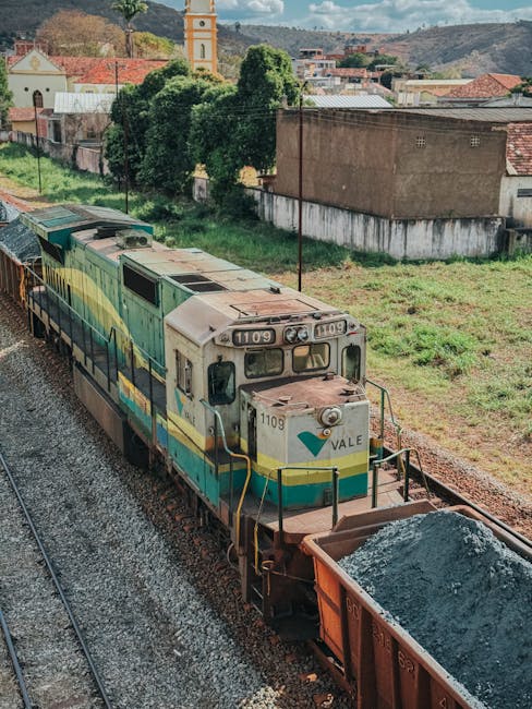 A diesel locomotive train with the number 1109 is visible on railway tracks, pulling a cargo wagon filled with loose gravel or stone, situated outdoors near a grassy area with patches of dirt. The train is painted in a combination of green, yellow, and white colors, and the locomotive features the name 'Vale' on its front. In the background, there are residential buildings with red-tiled roofs, a church tower with a yellow clock face, and various trees and greenery, indicating an urban environment adjacent to a suburban area. The lighting suggests a partly cloudy day with natural daylight illuminating the scene, which could be part of a logistical operation related to moving or transport services, similar to those provided by Man with Van Maida Vale.