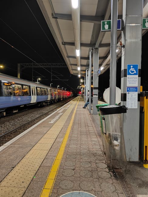 The image shows a view of Warwick Avenue station platform beneath an overhanging structure with a black and white striped canopy featuring decorative scalloped edging. A sign indicating the exit with an arrow and the words 'Way out' is visible, along with a smaller blue ‘Lift’ sign nearby. There are a few passengers waiting on the platform, some standing and others seated on a wooden bench, with dark clothing and backpacks. In the background, part of the train tracks, station signage for Golders Green, and urban buildings can be seen under a clear sky. The scene captures the typical setting for home relocation or furniture transport preparation, with potential for loading or unloading goods at the station for a house removal service, such as those offered by Man with Van Maida Vale.