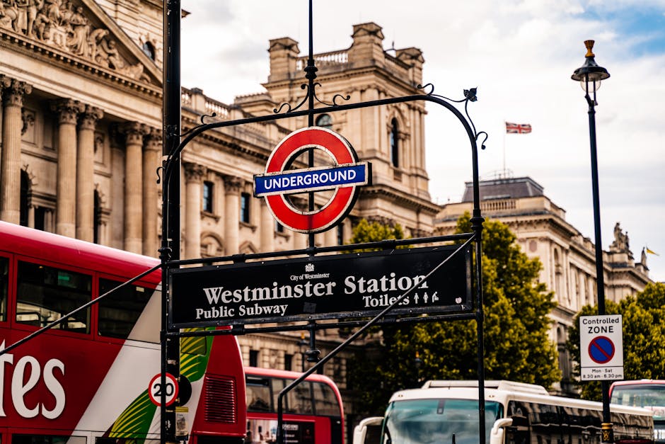 Close-up view of the Westminster Station underground sign mounted on a black wrought iron frame, with a red and blue London Underground roundel above it. The sign indicates the location of Westminster Station, a public subway station in London, with additional smaller text noting toilet facilities and staff access. Behind the sign, there is a historic building with detailed stonework and columns, typical of London architecture, and a flag flying from a flagpole on the roof. Part of a red double-decker bus is visible on the left side of the image, with its upper deck partially in view. To the right, a tall street lamp and a controlled zone parking sign are visible, along with trees and a glimpse of the skyline, under a partly cloudy sky. The scene captures an active urban environment in central London, relevant for contextualizing local transport hubs and cityscape in house removals and moving logistics, as managed by Man with Van Maida Vale.