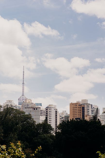 The image shows a cityscape with a clear sky and scattered clouds above a cluster of high-rise buildings and apartments. In the foreground, there are dark green trees and bushes, partially blocking the lower part of the scene. Prominent in the background is a tall, thin communication tower with antennas, rising above the other buildings. The buildings are made of brick, concrete, and glass, varying in height and design, suggesting a densely populated urban area. The scene is captured during daylight with natural lighting, illustrating an environment suitable for home relocation or furniture transport activities. This visual could relate to the logistics aspects of house removals, such as parking and access planning in urban settings, notably in areas like Maida Vale, as discussed on the 'westminster council rules Maida Vale parking for removals' page by Man with Van Maida Vale.